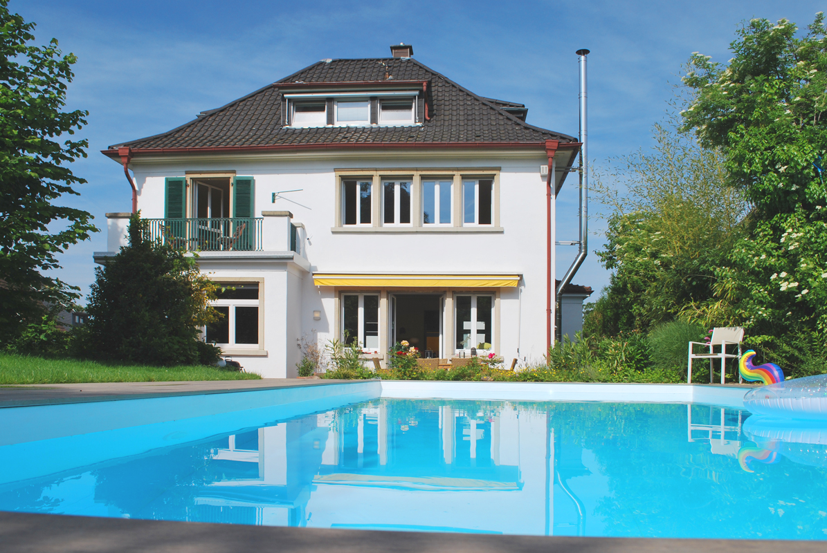 Aufnahme eines gepflegten Gartens mit eingebautem Pool im Sommer. Hinter dem Pool ist das Wohnhaus zu sehen.