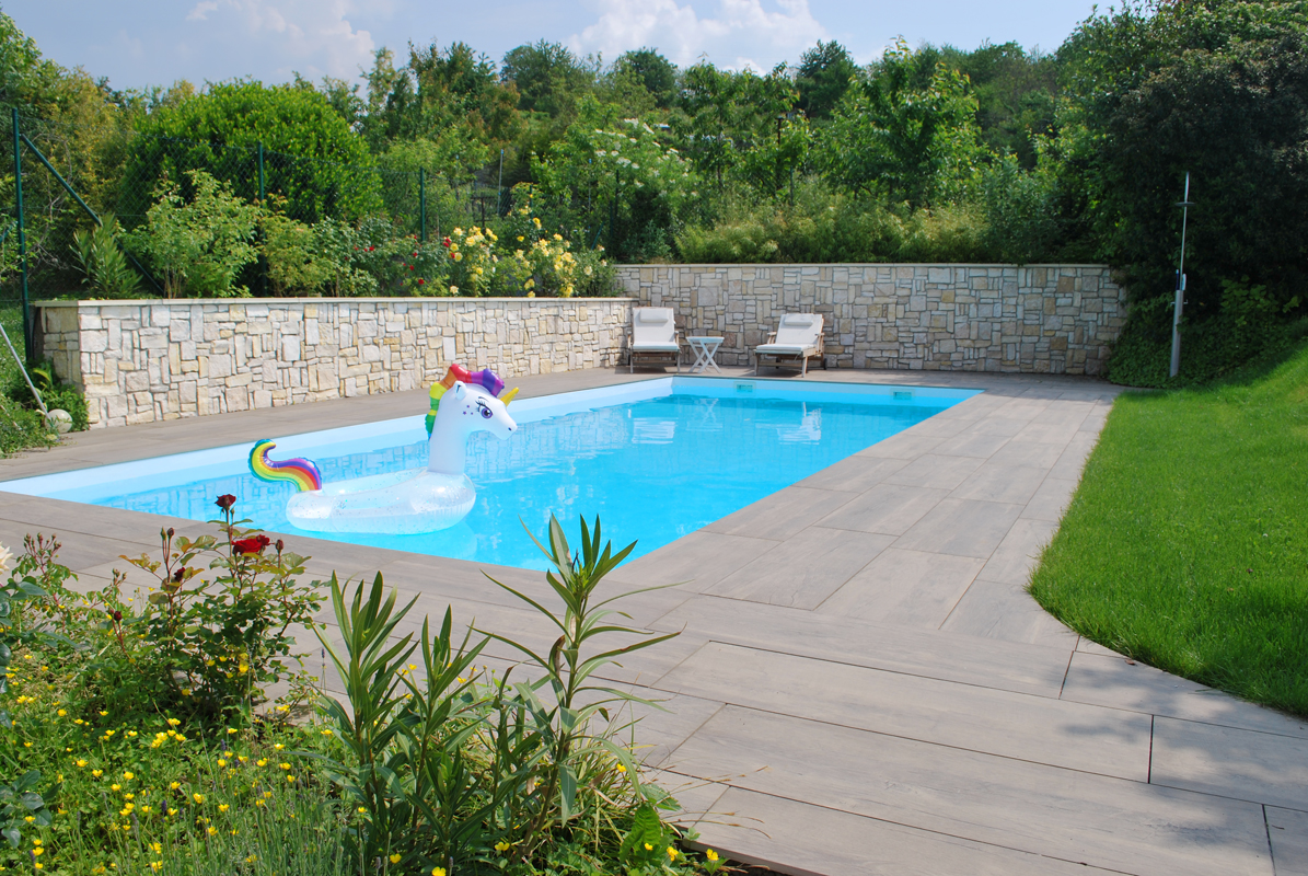 Aufnahme eines gepflegten Gartens mit eingebautem Pool im Sommer. Hinter dem Pool stehen moderne Liegen. Im Pool schwimmt ein Schwimmring in Einhornform.