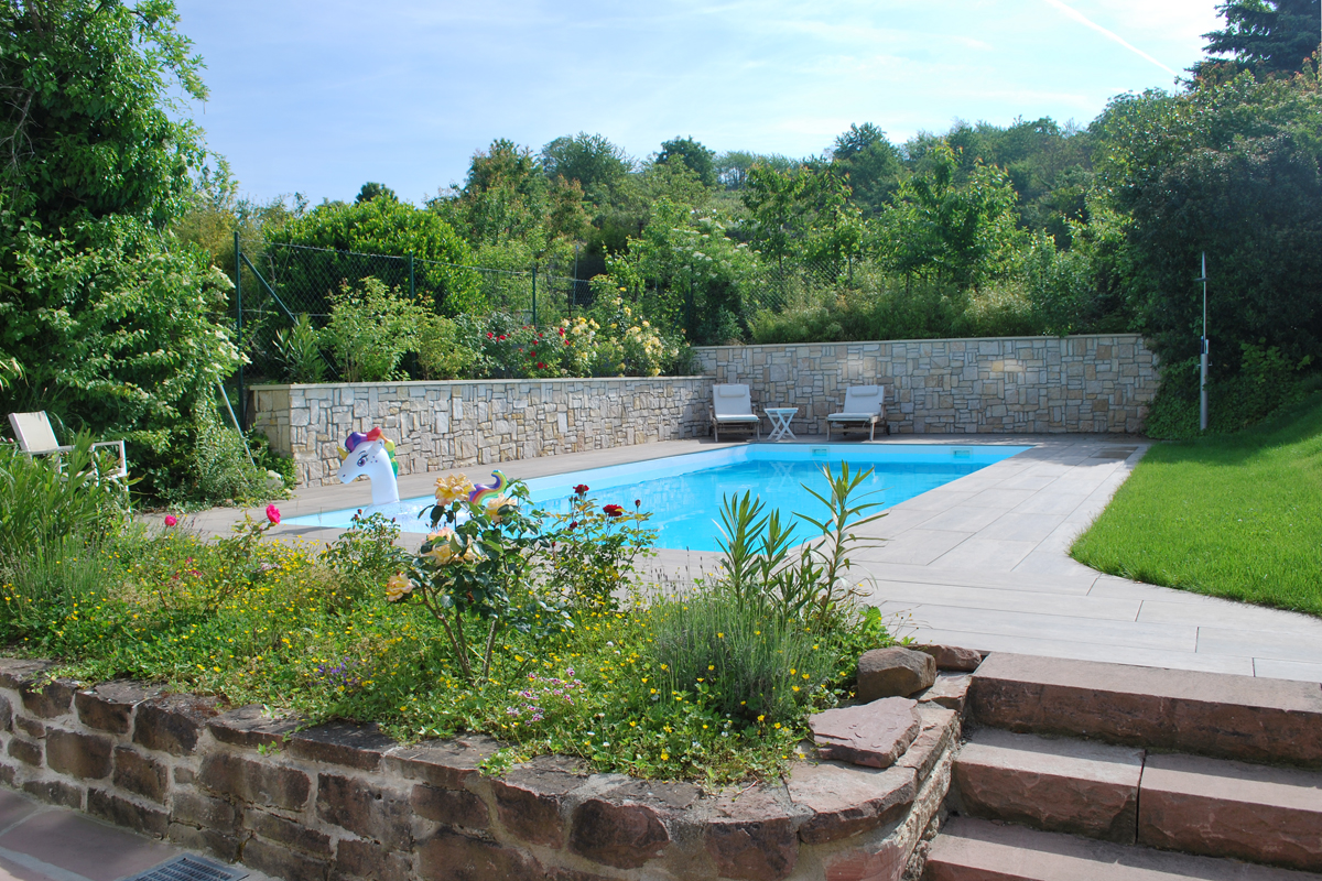 Aufnahme eines gepflegten Gartens mit eingebautem Pool im Sommer. Hinter dem Pool stehen moderne Liegen. Im Pool schwimmt ein Schwimmring in Einhornform.