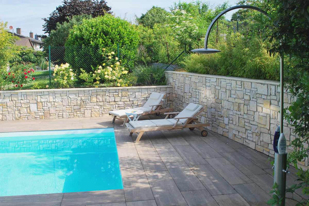 Neben einem eingebauten Pool in einem Garten stehen zwei moderne Holzliegen mit weißen Kissen. Davor steht eine eingebaute Outdoordusche.